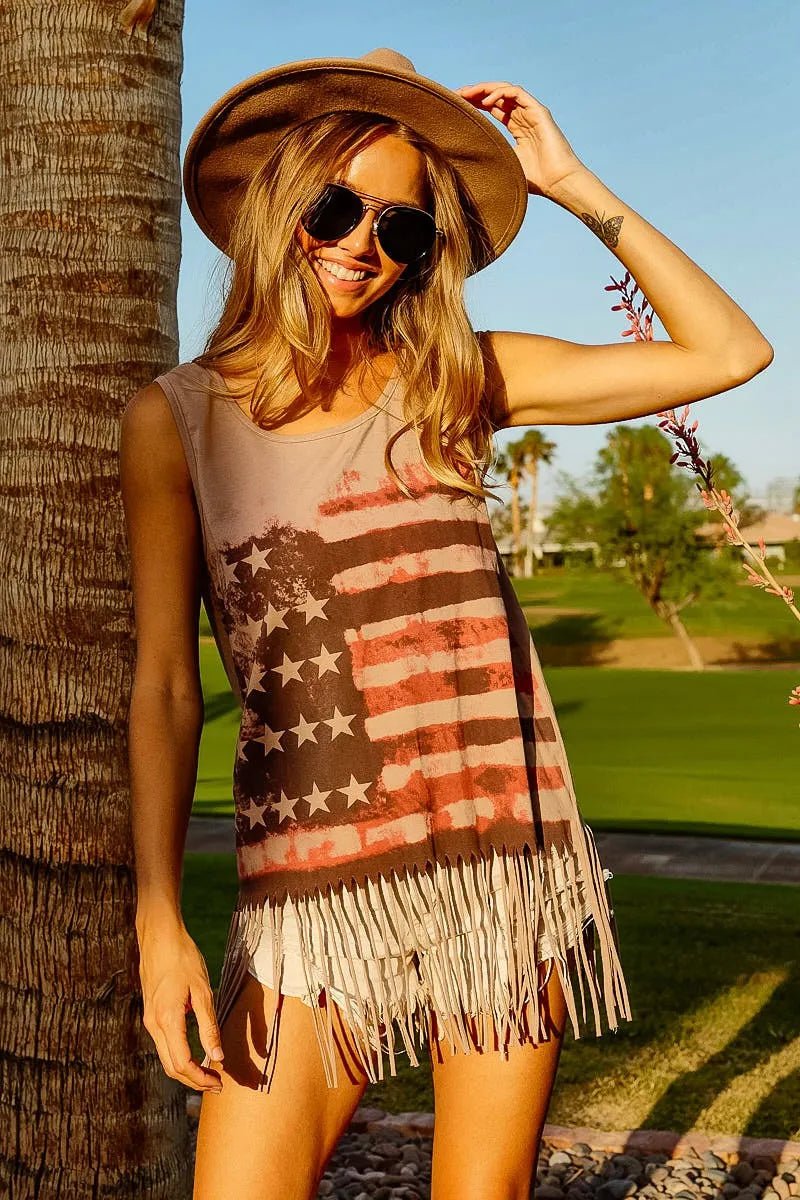 Woman wearing a sleeveless dress with an American flag design, sunglasses, and a hat, standing next to a palm tree.