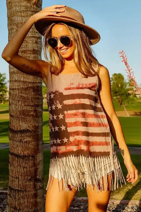 Woman wearing a fringe dress with an American flag design, sunglasses, and a hat in a sunlit outdoor setting.