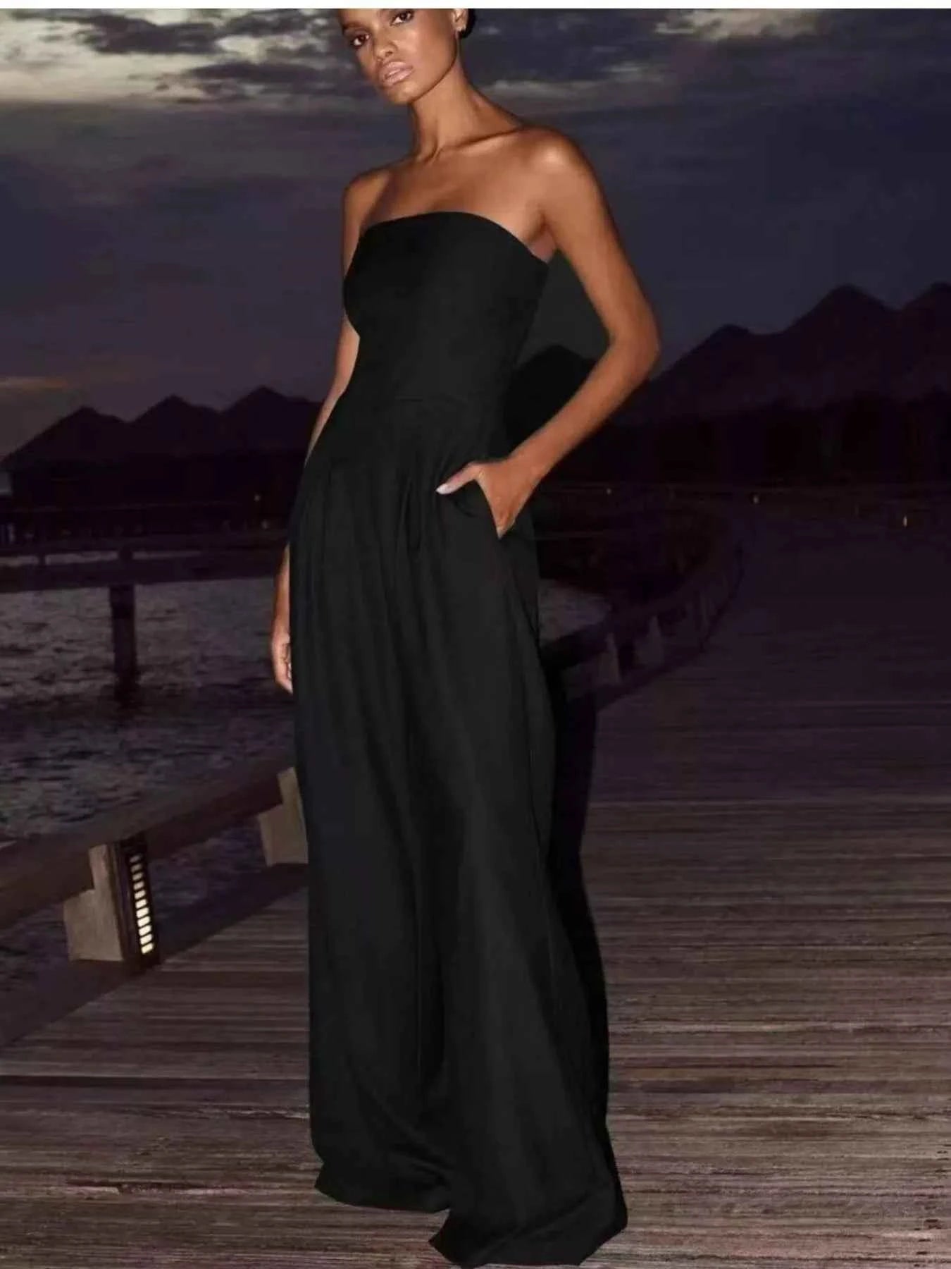 Woman in a black strapless gown standing on a dock with water and mountains in the background.