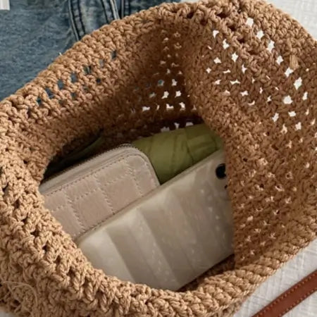 Close-up of a brown crochet bag with a green bottle inside, next to blue jeans on a white surface.