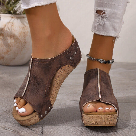 Brown open-toe sandals with cork wedge sole worn by a person on a wooden floor.