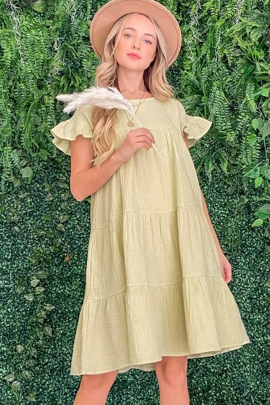 Woman in a light yellow dress and hat holding feathers against a green leafy background