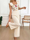 Woman wearing a floral embroidered top and white pants in a room with wooden floor and white walls.