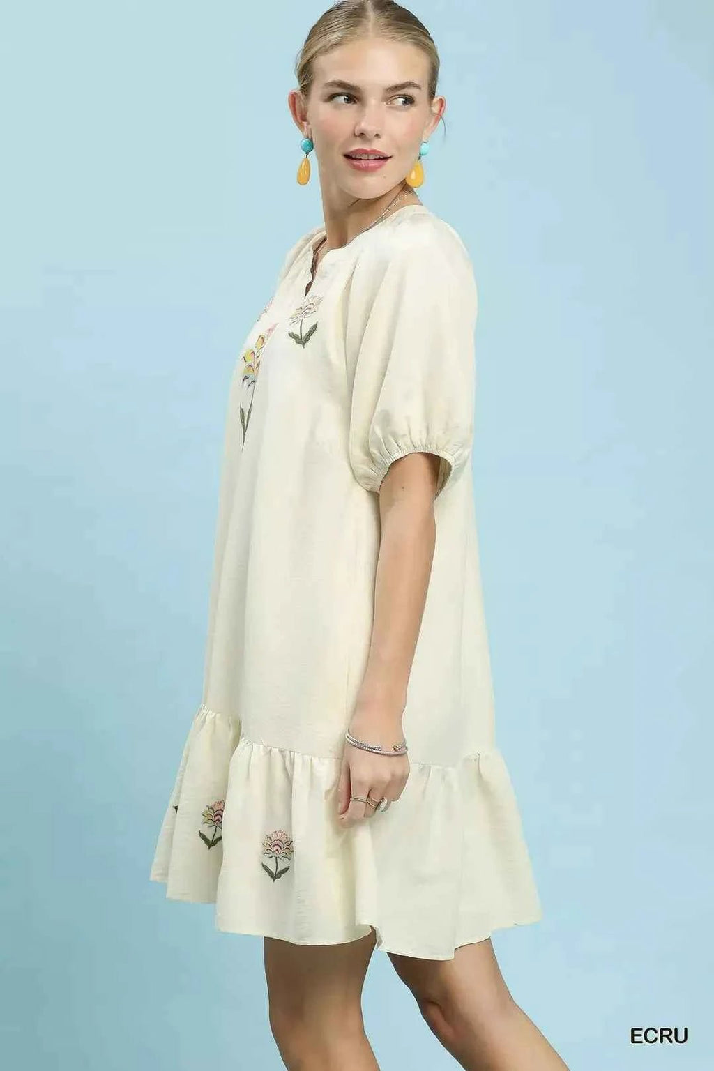 Woman wearing a cream-colored dress with floral embroidery on a light blue background glancing back