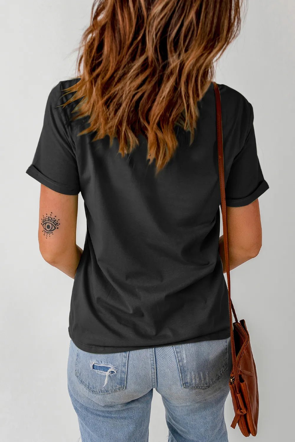 Person wearing a black t-shirt and blue jeans with a tattoo on their arm, standing against a plain background.