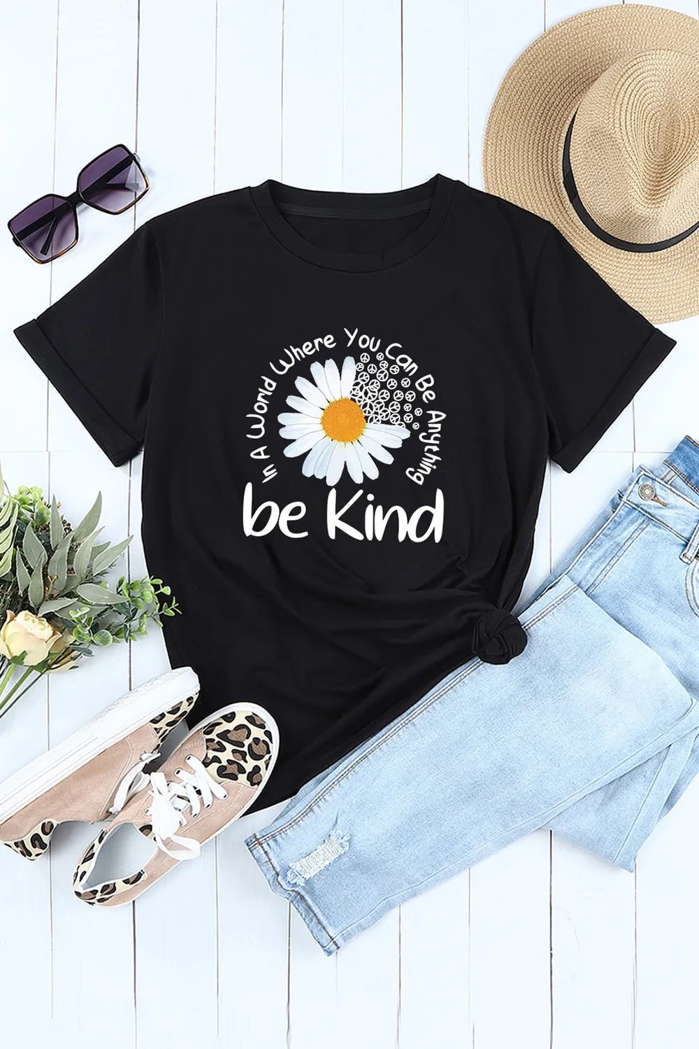 Black t-shirt with a flower graphic and text, placed on a white surface with sunglasses, hat, and jeans.