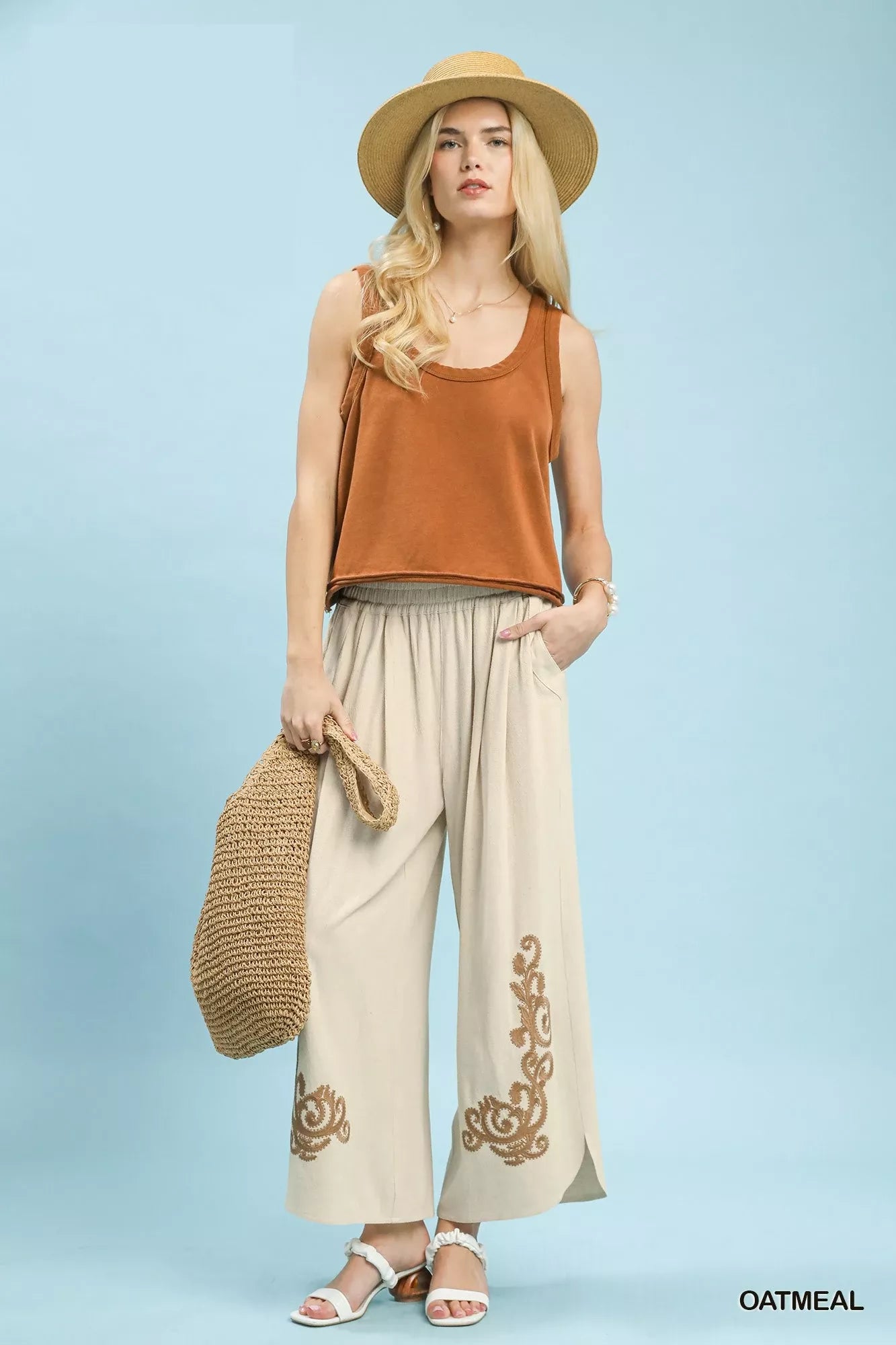 Woman wearing a rust-colored tank top and beige pants with embroidery, holding a straw bag against a light blue background.