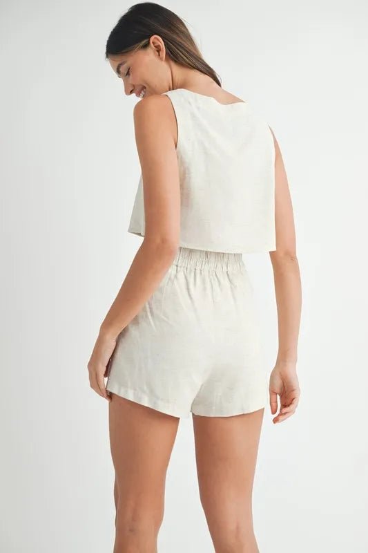 Woman wearing a white sleeveless top and shorts on a plain background turned around