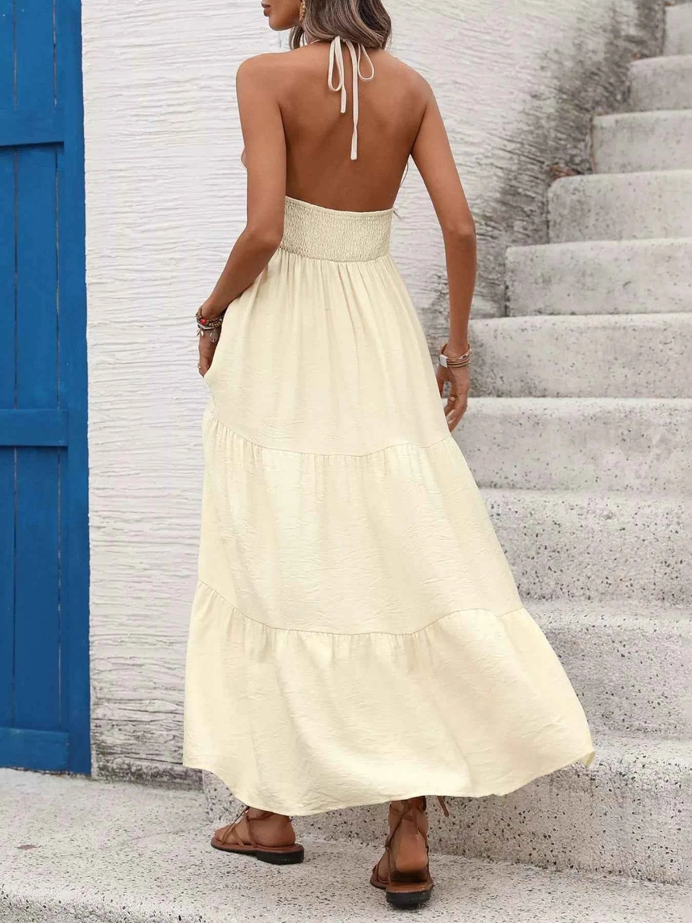 Woman in a long beige dress standing on stone steps with a blue door in the background turned away