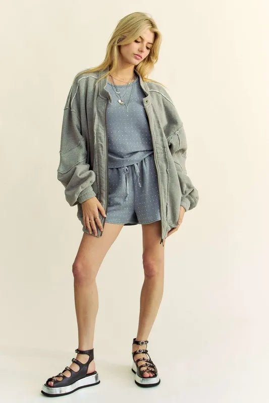 Woman wearing a gray outfit with a olive jacket and sandals on a plain background