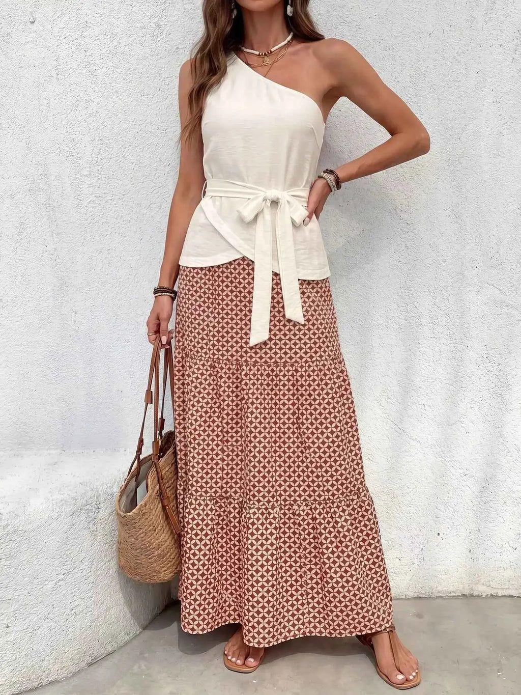 Woman wearing a white one-shoulder top and patterned skirt against a light gray wall.