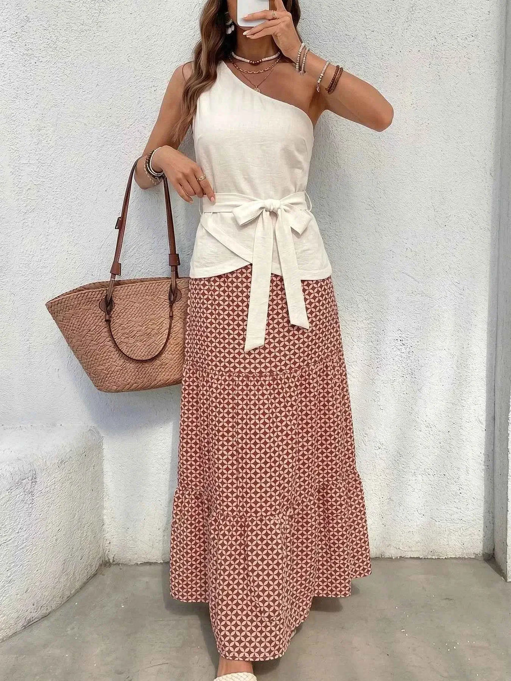 Woman wearing a white top and patterned skirt with a brown bag against a light gray wall.