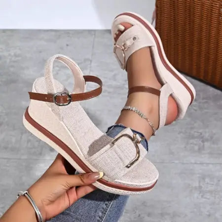 Beige wedge sandals with brown straps held by a hand on a gray floor.