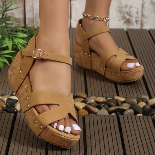 Tan sandals worn on a wooden deck with pebbles and plants in the background