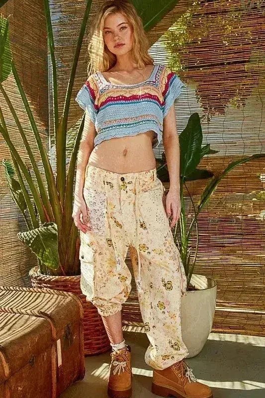 Woman wearing a colorful top and floral pants standing in a room with plants and wooden elements.
