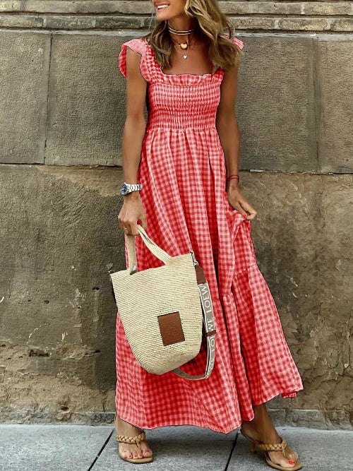 Woman in a red checkered dress holding a beige bag against a stone wall.