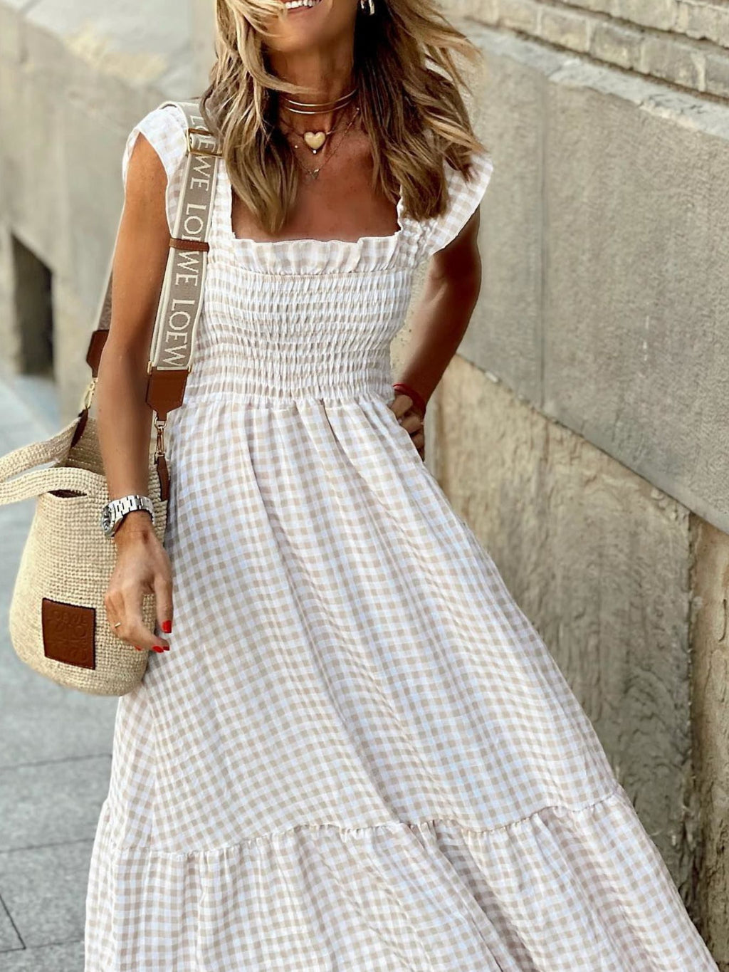 Woman wearing a white checkered dress with a straw bag against a stone wall.