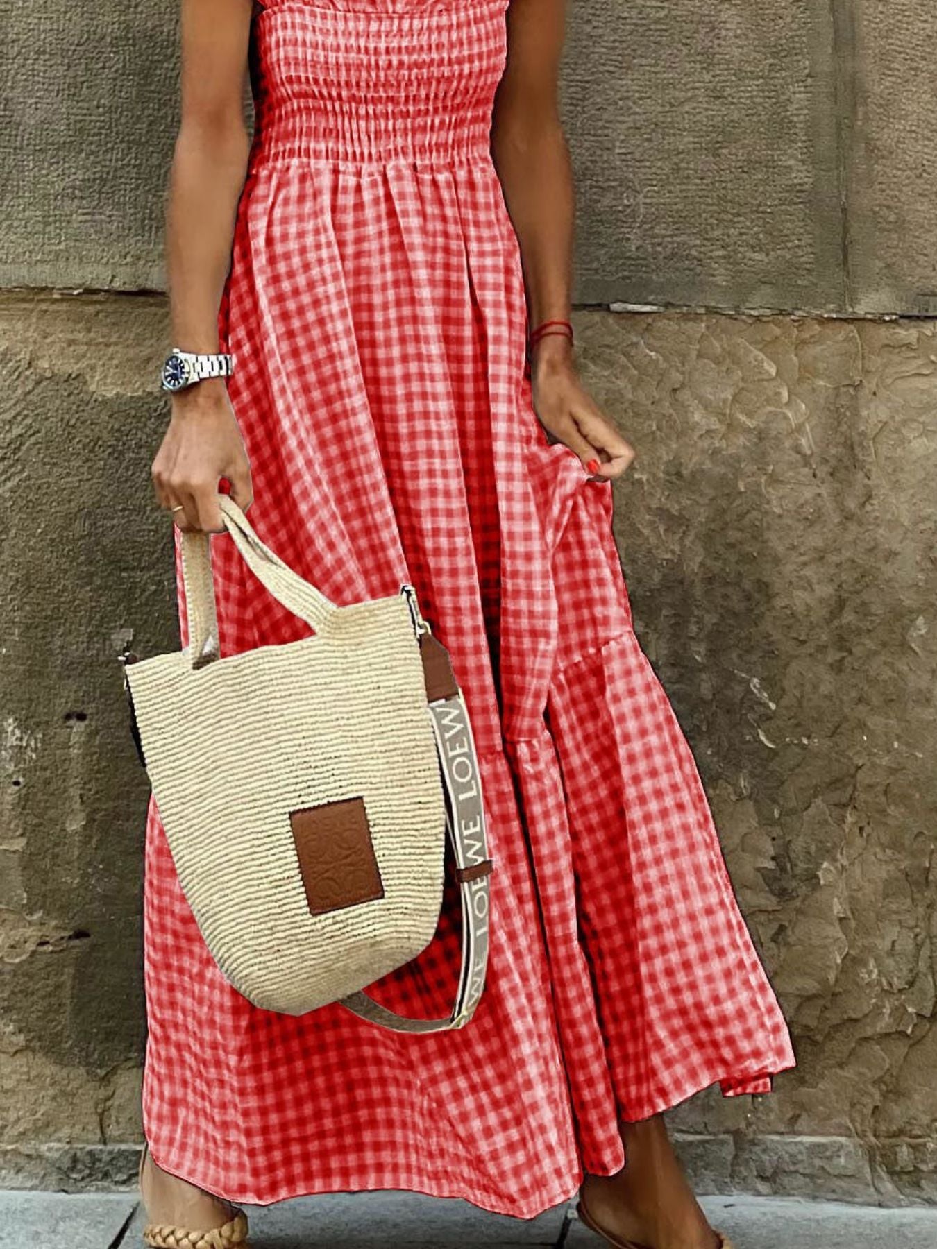 Person wearing a red checkered dress holding a straw bag against a stone wall.