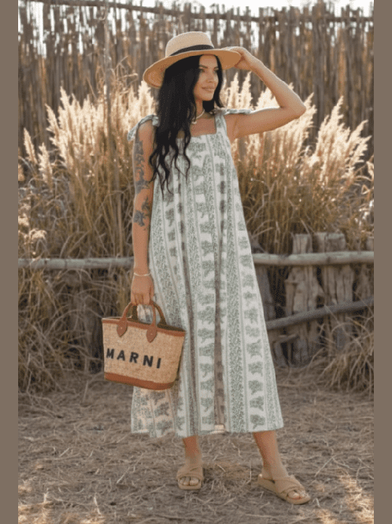 Woman in a patterned dress and straw hat standing in a natural setting with a wooden fence and tall grass.