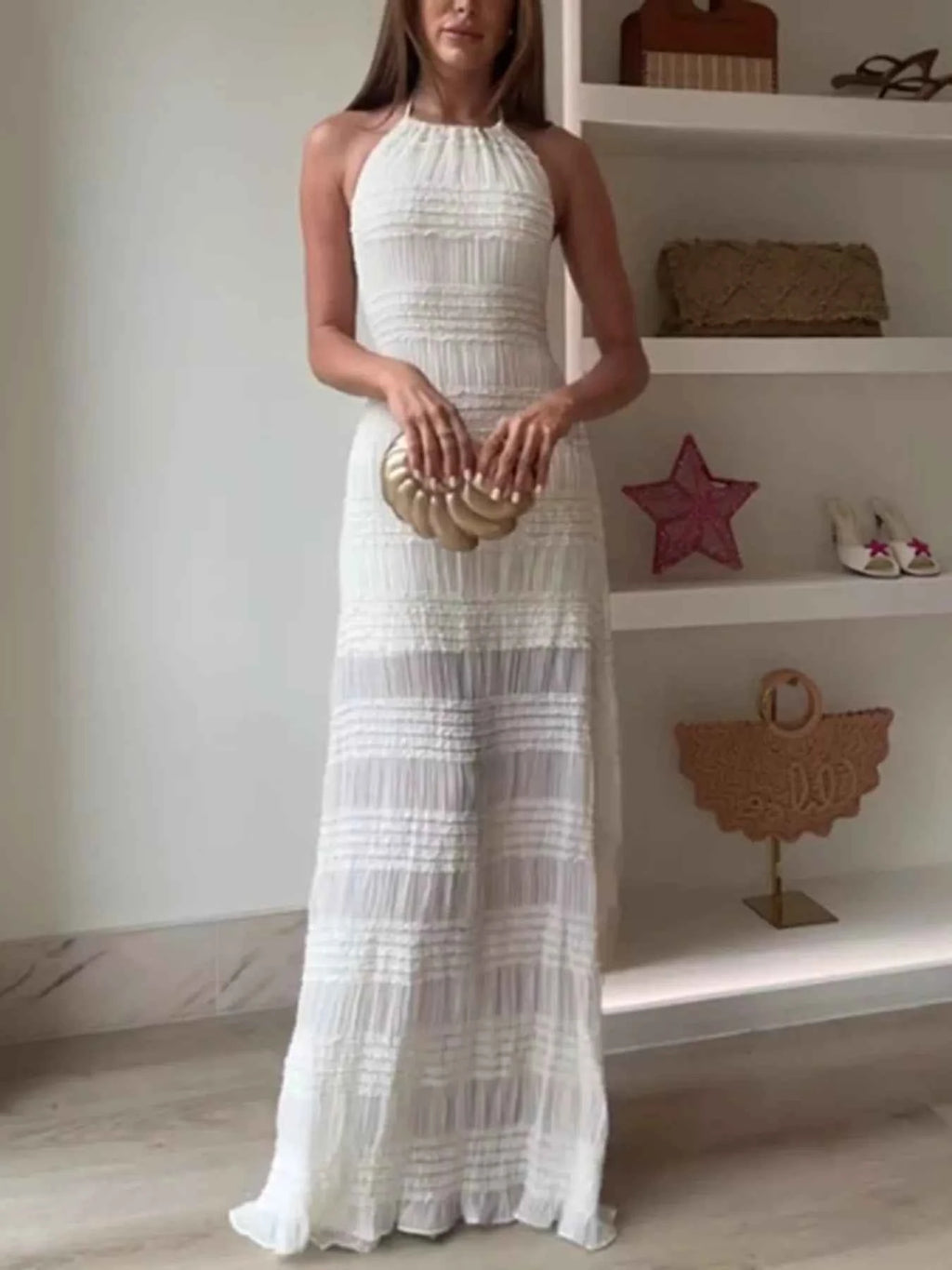 Woman in a white halter neck dress holding a gold clutch in a room with shelves.