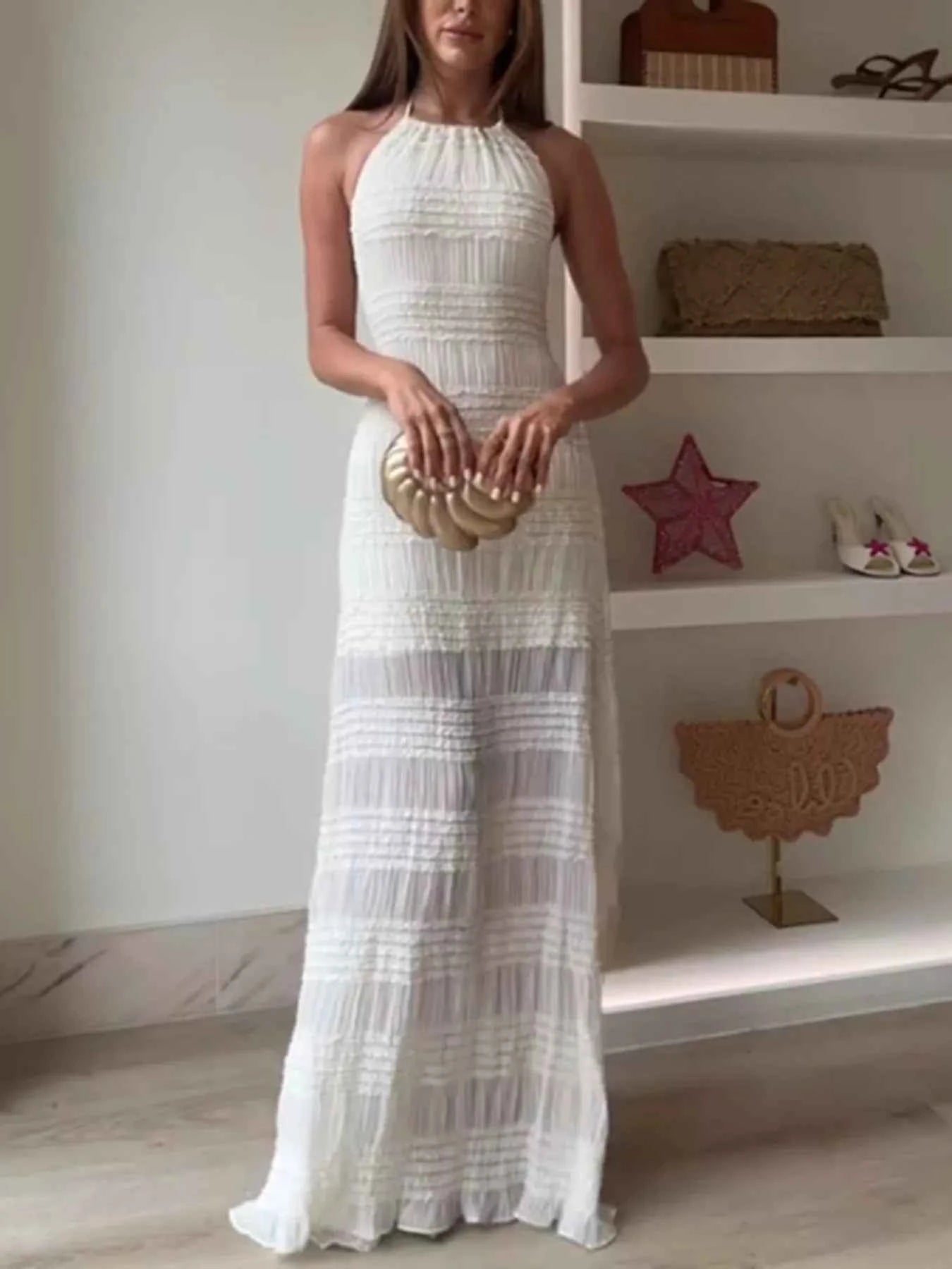 Woman in a white halter neck dress holding a gold clutch in a room with shelves.