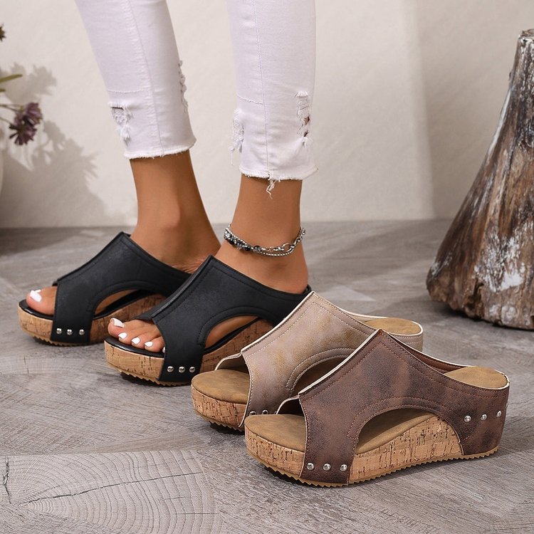 Black and brown open-toe sandals on feet with white pants, on a wooden floor.