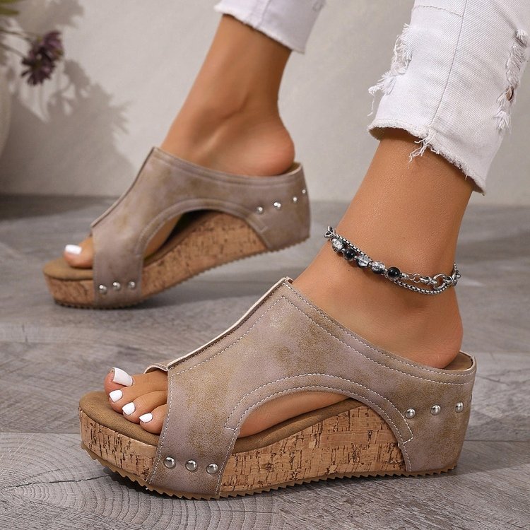 Beige wedge sandals with cork sole worn by a person on a gray floor.