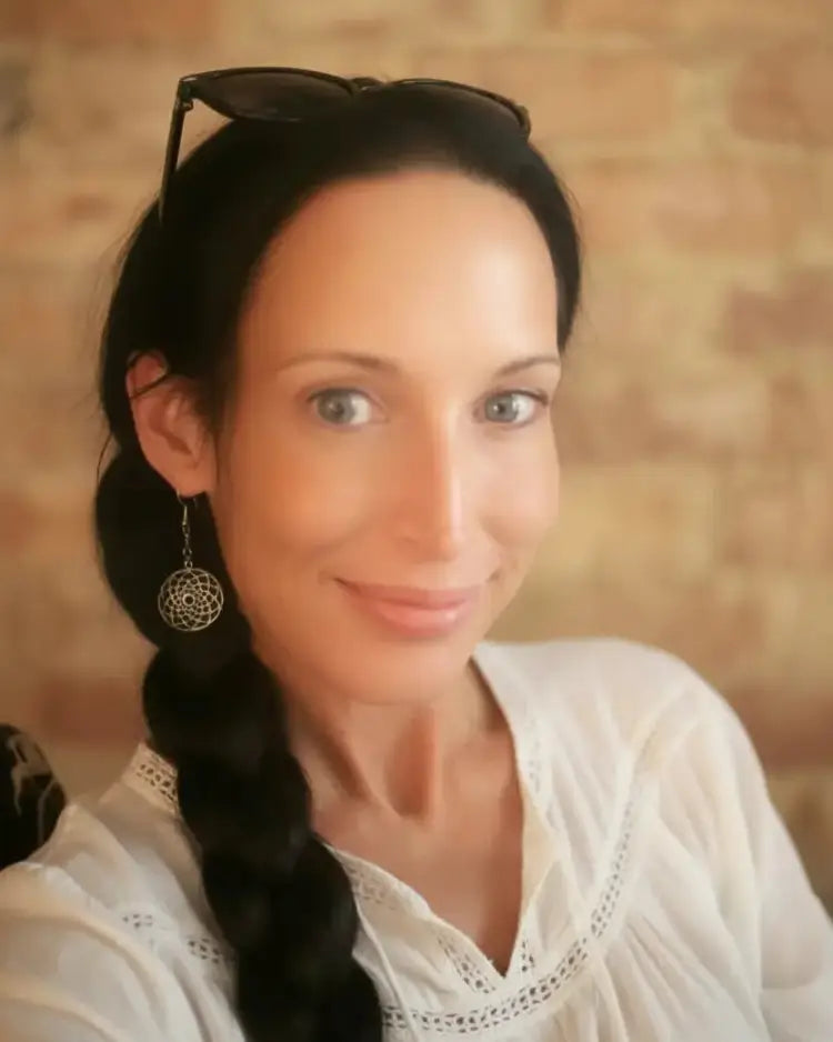 Woman with braided hair and sunglasses on head, wearing a white blouse against a brick wall.