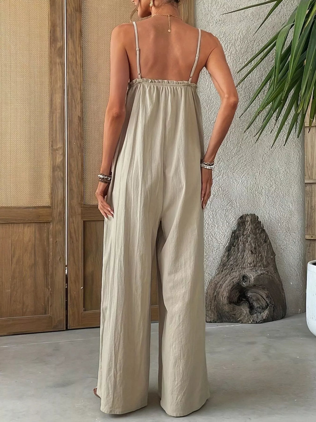 Woman wearing a beige jumpsuit standing in a room with wooden panels and a plant.