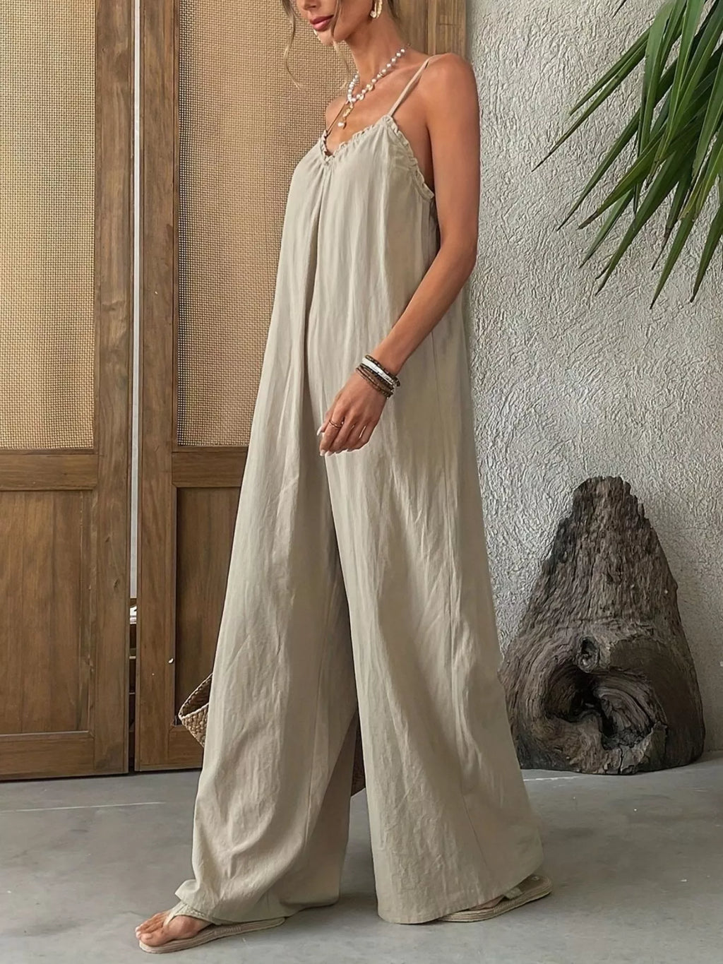 Woman wearing a light-colored jumpsuit standing against a neutral wall with a wooden door and plant in the background.