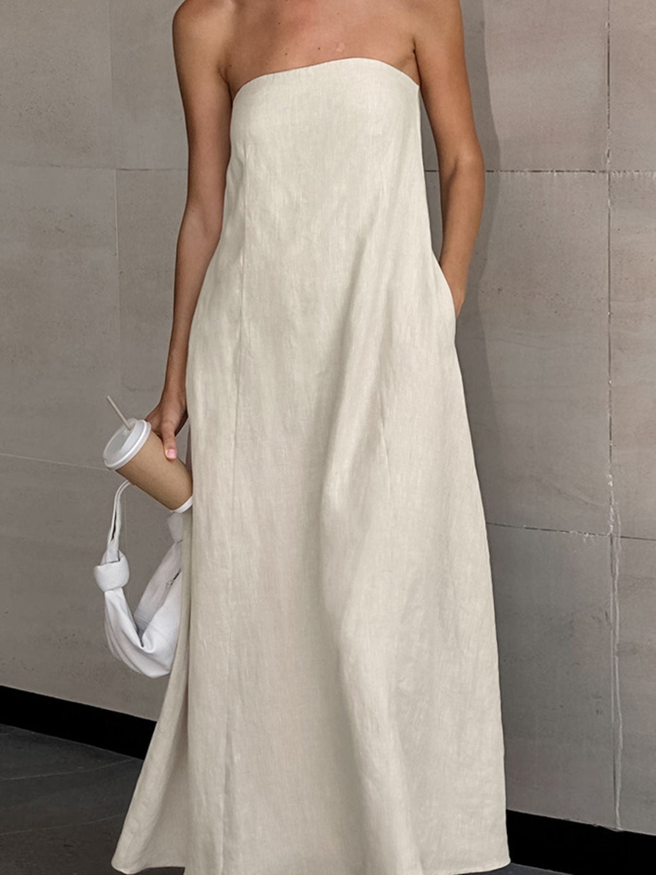 Smocked Back Tube Dress - Salty Season LLC Person wearing a long beige strapless dress holding a white bag and a coffee cup against a tiled wall.