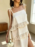 A woman wearing a cream-colored cotton slip dress with tie-shoulder straps and layered lace ruffles, holding a matching cream fabric bag in a sunlit indoor setting.