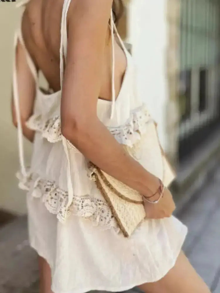 A woman wearing a cream-colored lace-trimmed mini dress and holding a woven straw clutch bag.