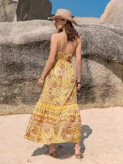 Woman in a yellow floral dress and wide-brimmed hat standing on a rocky beach.
