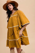 Woman wearing a mustard yellow dress with white trim and a brown hat on a beige background
