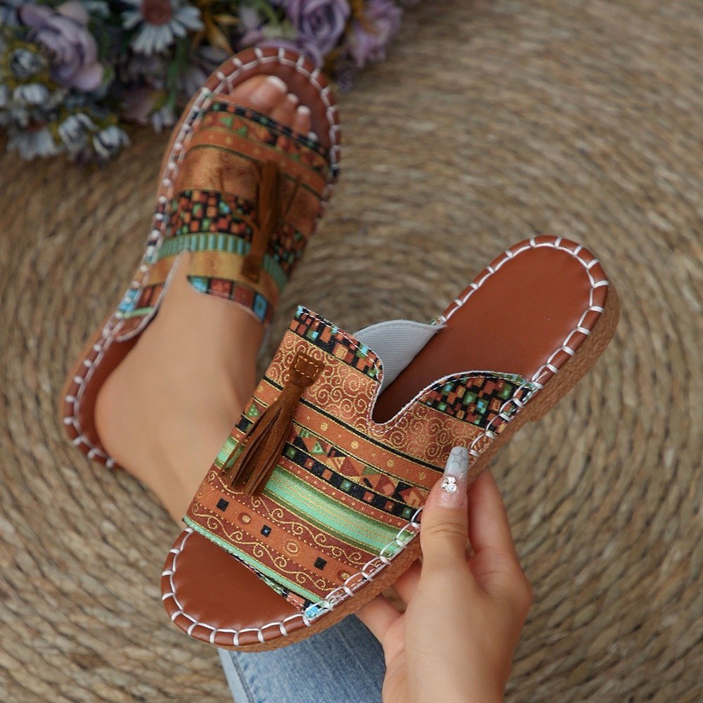 Hand holding a sandal with intricate patterns and a tassel on a woven surface.