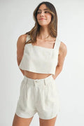 Woman wearing a white sleeveless top and shorts with hands behind back 
on a plain background