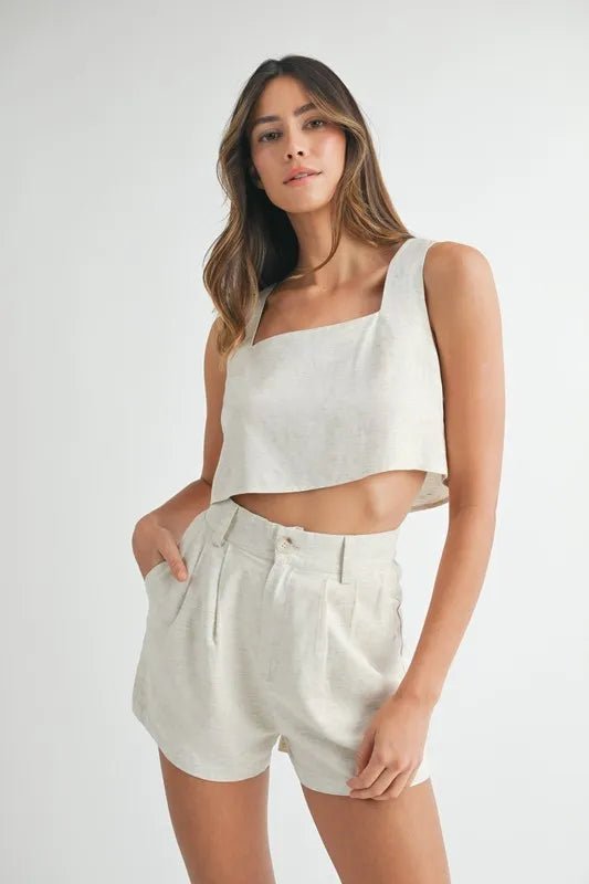 Woman wearing a white sleeveless top and beige shorts with right hand in pocket on a white background