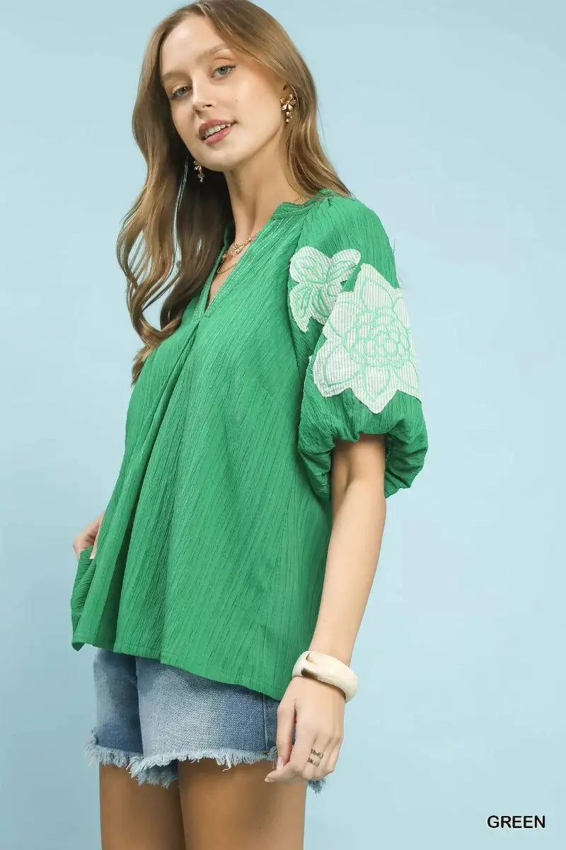 Woman wearing a green blouse with white floral embroidery on a light blue background
