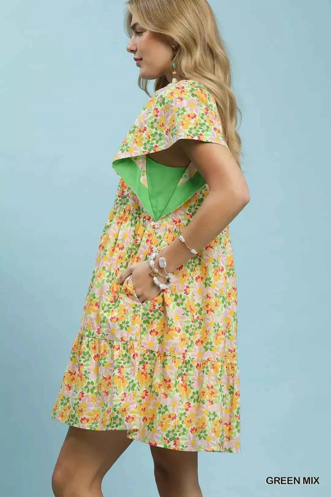 Woman wearing a floral dress with a green top against a light blue  standing sideways background
