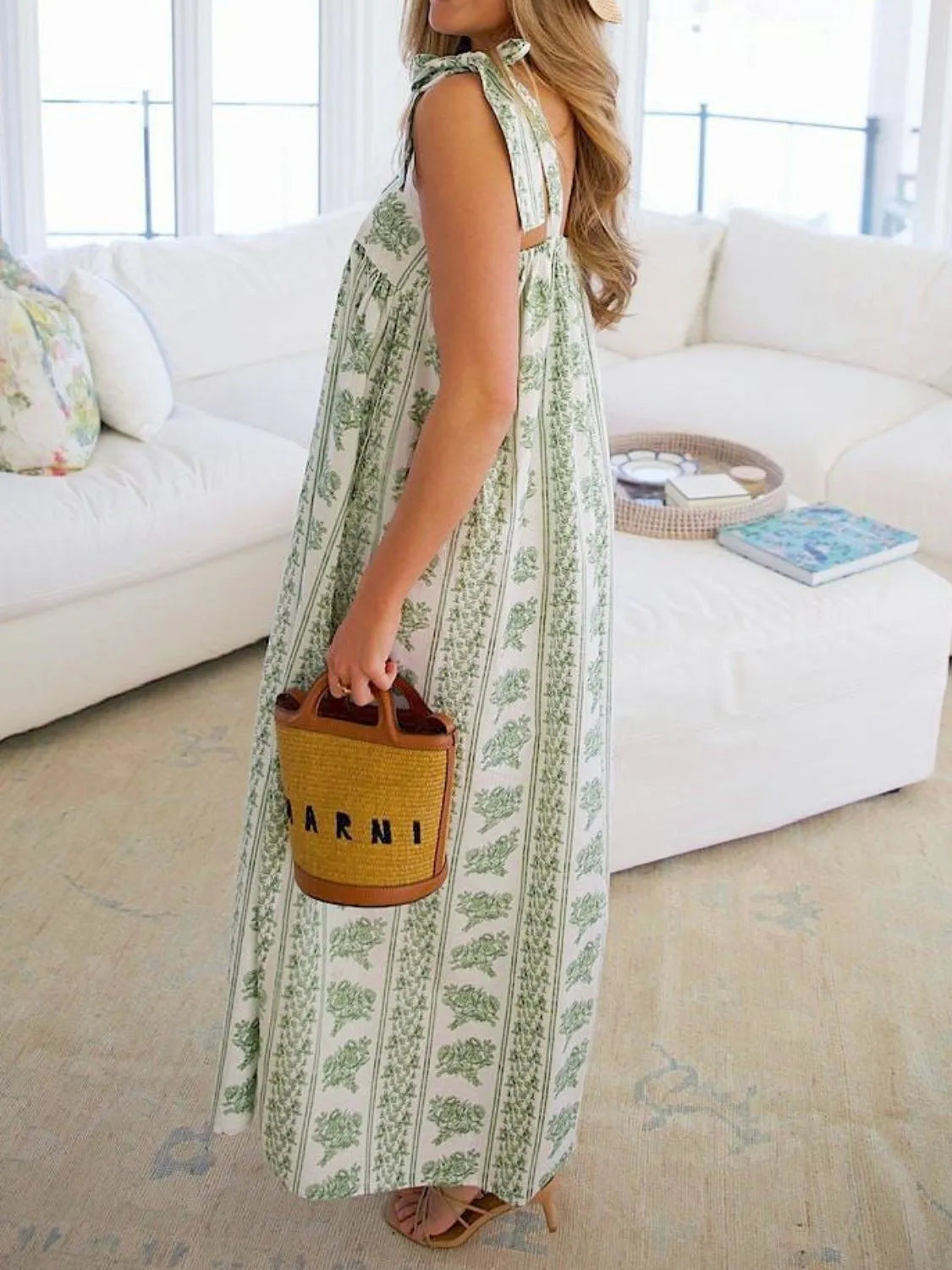 Woman in a long green and white patterned dress holding a brown bag with 'ARNI' branding in a living room.