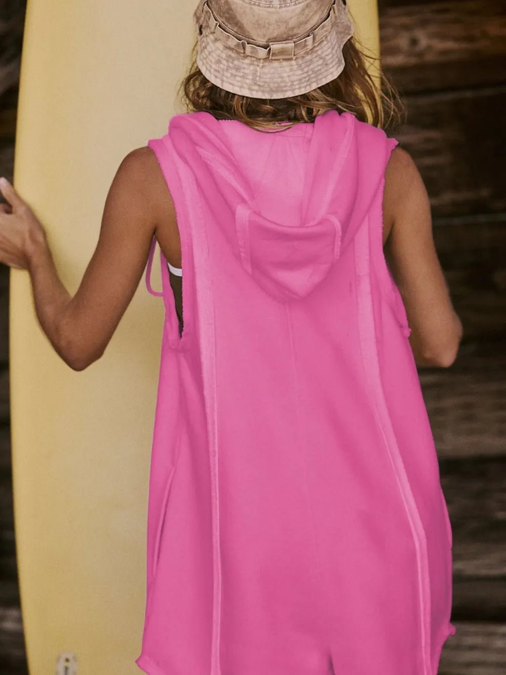 Person wearing a pink sleeveless hoodie with a hood, holding a yellow surfboard.