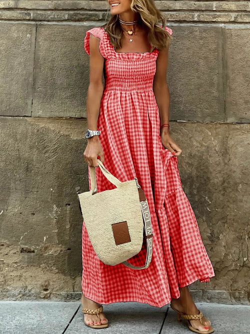 Woman in a red checkered dress holding a beige bag against a stone wall.