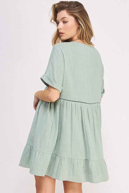 Blonde woman wearing a light green dress with ruffled layers on a white background turned towards wall